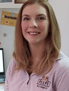 Lis is wearing a lilac polo shirt that has the Peeps' logo on it. It's a headshot and she is smiling. There's a postcard with #HeardOfHIE in the background.