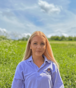 A lady with long blonde hair wearing a lilac t-shirt with the Peeps' logo on. She is smiling. The background shows grass and blue skies.