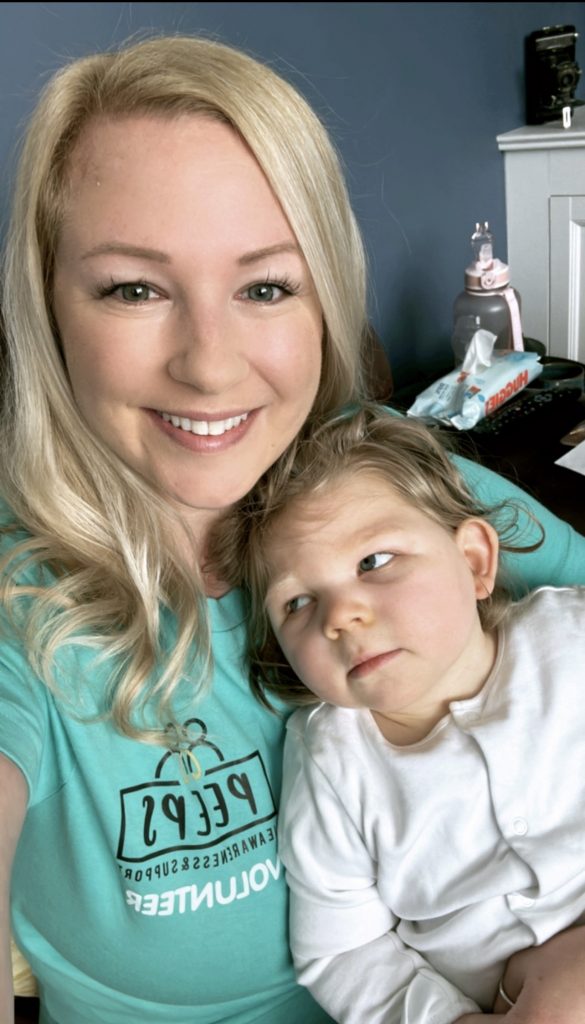 Mum Jen is holding her daughter Ada as she smiles at the camera. Jen has blonde hair and is wearing a turquoise t-shirt with the Peeps logo volunteer on. Ada is wearing white baby grow.