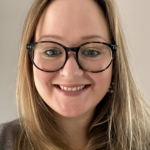 A headshot of a lady smiling at the camera. She has long hair and is wearing glasses.