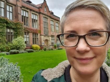 Sarah standing in front of a large building. Sarah has short hair and dark framed glasses.
